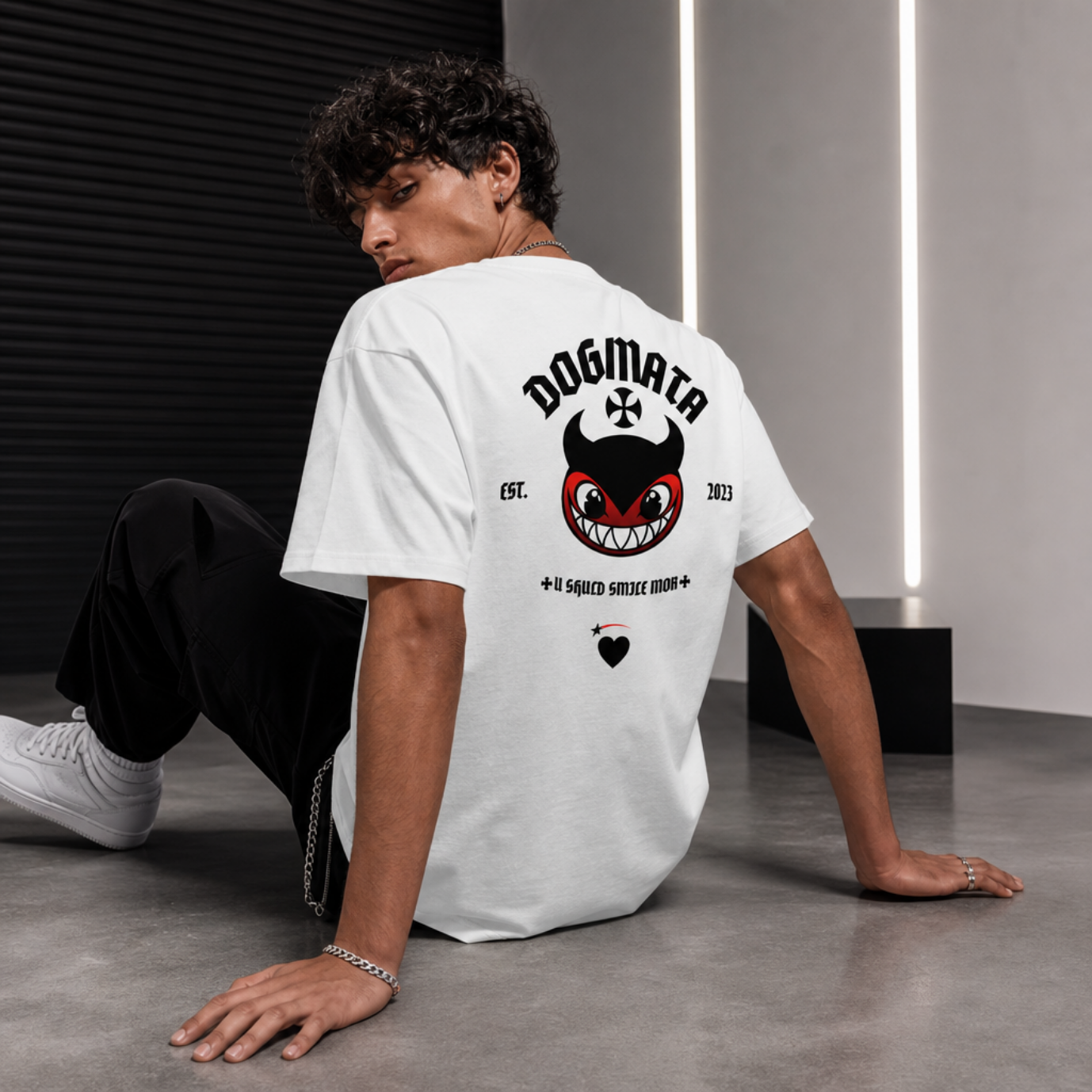 Model wearing a white t-shirt with a graphic design on the back, sitting on a gray floor.