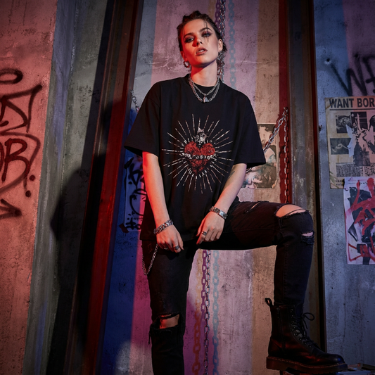 A model wearing a black graphic t-shirt with a red and silver heart design, paired with ripped black jeans and black boots, posing in front of a graffiti-covered concrete wall.