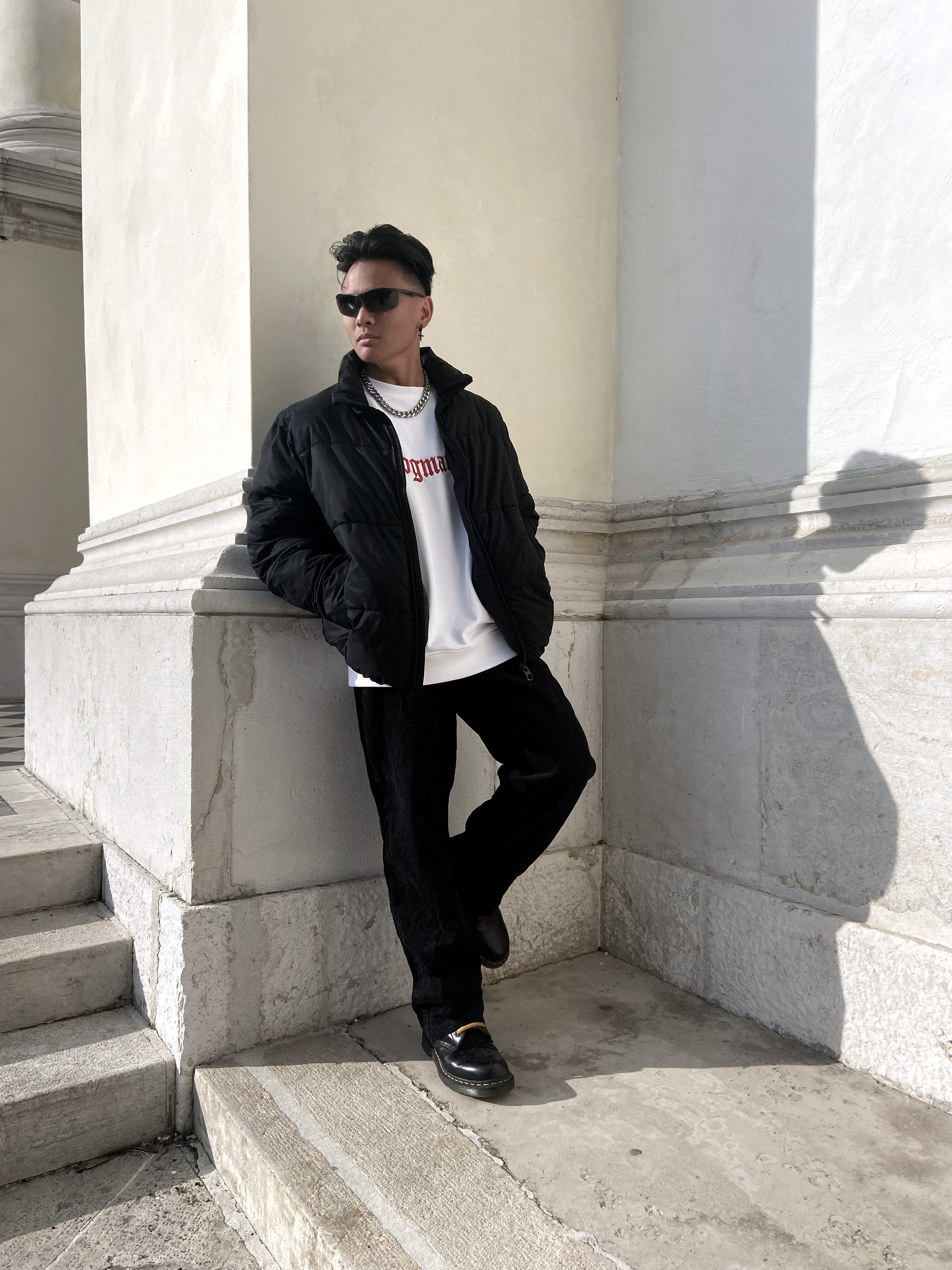 Model wearing a black jacket with a white organic cotton sweatshirt and black pants, standing on steps with hands in their pockets.